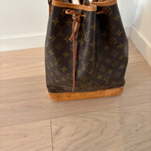Louis Vuitton väska - Säljer en klassisk och den ikoniska  Noe Monogram från Louis Vuitton med det ikoniska monogrammönstret. Väskan har läderdetaljer och en praktisk dragskoöppning. Perfekt för dig som vill ha en tidlös och stilren accessoar. Äkthetsintyg följer givetvis med. 