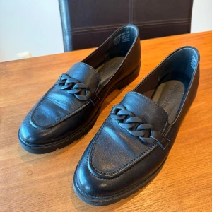 Svarta skinn loafers från Tamaris - Snygga svarta loafers från Tamaris med en dekorativ kedjedetalj på ovansidan. Skorna är tillverkade i skinn och har en robust sula för extra komfort. Perfekta för en stilren look.