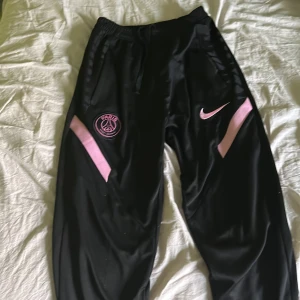 Svarta träningsbyxor från Nike - Snygga svarta träningsbyxor från Nike med rosa detaljer och PSG-logga. Byxorna har elastisk midja med snörning och sidofickor. Perfekta för sport och avslappnade dagar.