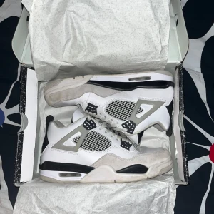 Nike Air Jordans 4 military black - Snygga Nike Air Jordans i vitt och grått med svarta detaljer. Stilrena och snygga. Skorna är inte så mycket använda men har fått en defekt som du kan se på bild 5, säljer för att jag inte använder dom längre, är i storlek 45 men skulle säga passar 44