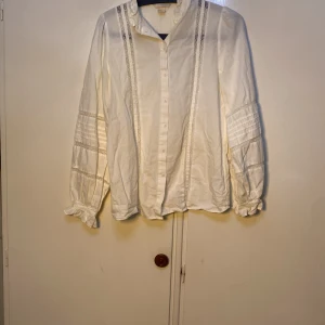 Vit blus från H&M - Elegant vit blus från H&M med långa ärmar och fina spetsdetaljer längs framsidan. Perfekt för en stilren look. Blusen har knappar framtill och en klassisk krage.