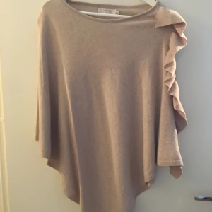 Beige tröja med volangdetalj - Säljer en stilren beige tröja poncho med unik volangdetalj på ena axeln. Tröjan har en lös passform och trekvartsärmar, perfekt för en avslappnad look. Passar bra till både jeans och kjol.