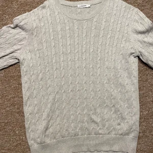 Grå stickad tröja från j.lindeberg - Säljer en stilren grå stickad tröja från j.lindebeeg med kabelstickat mönster. Tröjan har rund hals och långa ärmar, perfekt för kyligare dagar. Så god som ny använt fåtals gånger
