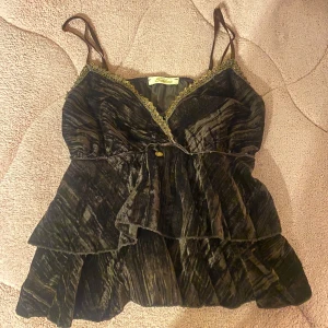 Volang topp brun guld - Jättefin volang topp från märket Sashoo som är i samett material och brun med guldiga detaljer. Står ingen storlek men skulle säga att den passar S/M❣️ Säljer billigare då ena bandet är trasigt och längden inte går att justera på det bandet (se bild 4)