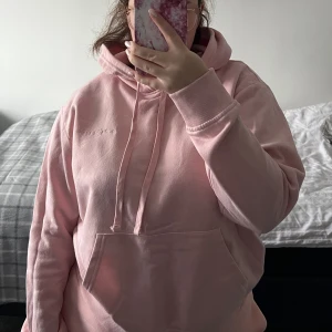 Rosa hoodie med broderad text - Mysig rosa hoodie med broderad text på bröstet. Den har en stor ficka framtill och en justerbar huva. Den är i mycket bra skick, endast använd ett par gånger