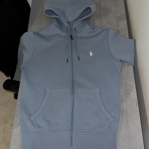 Baby Blå hoodie från Ralph Lauren - Säljer en snygg blå hoodie från Ralph Lauren med ärmar och dragkedja. Den har en klassisk logga på bröstet och en praktisk huva med dragsko. Perfekt för en avslappnad stil.