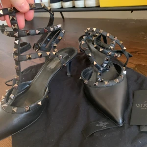 Svarta Valentino Rockstud klackskor - Snygga svarta klackskor från Valentino med spetsig tå och ikoniska Rockstud-detaljer. Skorna har remmar dekorerade med nitar som ger en edgy look. Dom är i väldigt fint skick och klackarna är 5cm. Storlek 39
