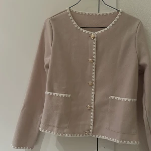 Beige kofta med dekorativa knappar - Elegant beige kofta med dekorativa knappar och vita detaljer längs kanterna. Den har två praktiska fickor framtill och långa ärmar. Perfekt för en stilren look. Nytt skick.