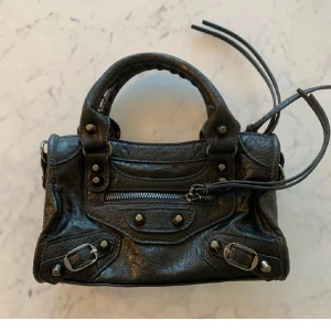 Svart axelväska med nitar - Snygg svart axelväska i skinn med coola nitar och dragkedja. Väskan har dubbla handtag och en avtagbar axelrem. Perfekt för att ge din outfit en edgy touch.