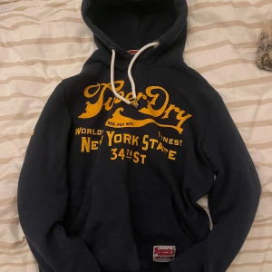 Mörkblå hoodie från Superdry - Säljer en mörkblå hoodie från Superdry med gul text på framsidan och siffran 34 på ärmen. Den har en klassisk känguruficka och justerbar huva med vita snören. 