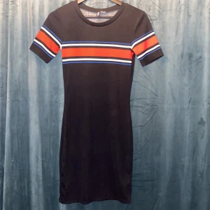 Svart klänning från H&M Divided - Snygg svart klänning från H&M Divided med korta ärmar och färgglada ränder i rött, blått och vitt över bröstet. Perfekt för en avslappnad stil med en touch av färg. Klänningen har en normal passform och är tillverkad i ett mjukt material.
