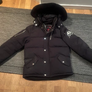 Moose knuckles  - Snygg svart dunjacka med avtagbar huva och pälsdetaljer. Jackan har dragkedja och knappar framtill samt praktiska fickor. Perfekt för kalla vinterdagar. Säljer jackan eftersom den inte passar. Är strl XL i barn men passar bättre som XS vuxen.