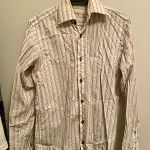 Vit och brunblå randig herrskjorta från Batistini i stl 35/36 - Använd fåtal ggr, i fint skick. Snygg vit skjorta från Batistini med bruna och blå ränder. Skjortan har långa ärmar och knappar framtill. Perfekt för en stilren look.