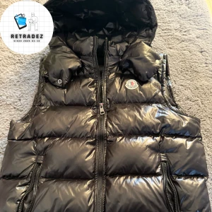 Moncler Väst - Snygg svart dunväst från Moncler med glansig finish och dragkedja framtill. Västen har en praktisk huva och Monclers logotyp på bröstet. Perfekt för kyligare dagar.