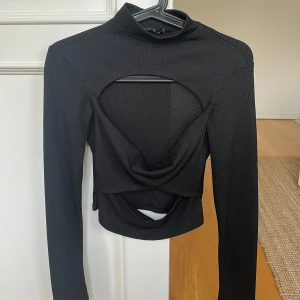 Svart ribbad topp med öppen design - Snygg svart ribbad topp med långärmad design och unik öppen detalj framtill. I gott skick🖤