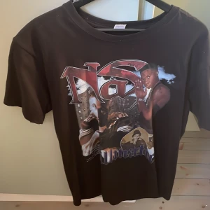 Svart t-shirt med Nas-tryck - Cool svart t-shirt med stort tryck av Nas på framsidan. Perfekt för fans av hiphop och streetwear. T-shirten har en avslappnad passform och korta ärmar.