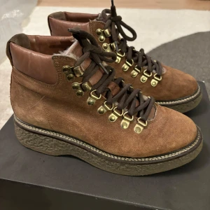 Rizzo Bruna mockaboots med snörning - Rizzo Bruna mockaboots med snörning. Snygga bruna boots i mocka med snörning och guldfärgade detaljer. Skorna har en robust sula och är fodrade för extra värme. Perfekta för kyligare dagar.