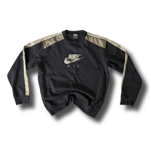 Svart och beige oversize tröja från Nike - 📏: L 64cm / B 52cm | Snygg svart tröja från Nike med beige detaljer på ärmarna och broderad logga framtill. Perfekt för en sportig look. Tröjan har långa ärmar och en bekväm passform.