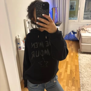 Svart hoodie med franska glittrande text - Säljer en svart hoodie med glittrande texttryck på franska och stjärna på framsidan. Den har en klassisk design med huva och långa ärmar. Perfekt för en avslappnad stil. Strl 164 passar xs och s🩷
