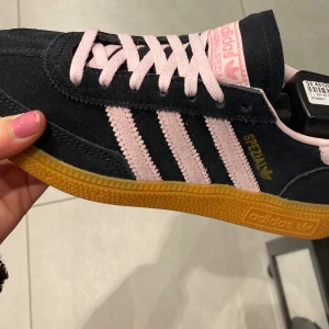 Adidas Gazelle i svart och rosa - Snygga Adidas Gazelle sneakers i svart mocka med rosa detaljer och snörning. Skorna har en klassisk design med gummisula och de ikoniska tre ränderna i rosa på sidan. Perfekta för en stilren look.