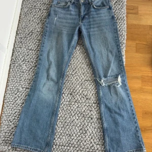 Blå bootcut jeans - Snygga blå bootcut jeans med slitningar på ena benet. Klassisk femficksdesign och knappgylf. Perfekta för en avslappnad stil.