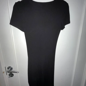 Svart klänning med rynkdetaljer - Snygg svart klänning med rynkdetaljer längs sidan. Perfekt för en stilren look. Klänningen har korta ärmar och en figurnära passform.