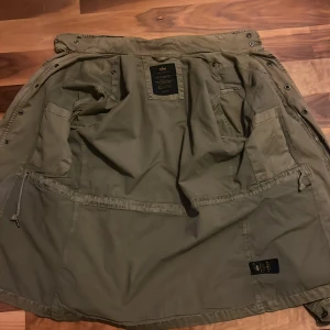 Olivgrön jacka från Alpha Industries - Säljer en olivgrön jacka från Alpha Industries. Jackan har flera fickor och en cool detalj med en röd tagg på ärmen. Perfekt för höst och vår med sin robusta design och praktiska knappar.