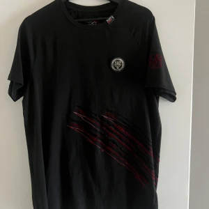 Svart t-shirt från Plein Sport - Snygg svart t-shirt från Plein Sport med coola röda detaljer och ett märke på bröstet. T-shirten har korta ärmar och ett tryck med siffran 78 på baksidan. Perfekt för en sportig look!