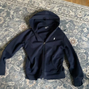 Mörkblå hoodie med dragkedja - Säljer en stilren mörkblå hoodie från Ralph Lauren med dragkedja och huva. Perfekt för en avslappnad look. Den har långa ärmar och en liten logotyp på bröstet. Passar bra till både jeans och träningsbyxor. Pris kan diskuteras!