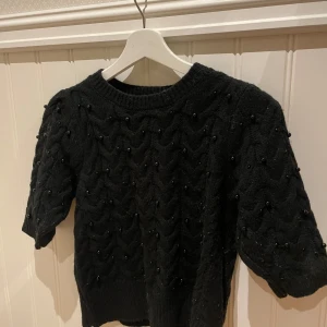 Svart stickad tröja från Zara - Säljer en svart stickad tröja från Zara med korta ärmar och ett snyggt mönster. Tröjan har små pärlor som detaljer och en rund halsringning. Perfekt för att ge din outfit en elegant touch.