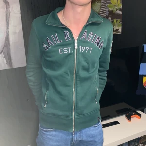 Grön hoodie från Sail Racing - Säljer en grön hoodie från Sail Racing med dragkedja och tryck på bröstet. Tröjan har långa ärmar och en patch på ärmen. Perfekt för en avslappnad stil. Modellen är 180 cm och väger 60 kg 