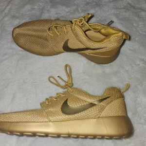 Guldiga sneakers från Nike - Snygga guldiga sneakers från Nike med snörning och en stilren design. Perfekta för dig som vill sticka ut med en unik färg. Skorna har en bekväm passform och är tillverkade i syntetmaterial.