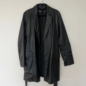 Svart skinnjacka från Zara - Snygg svart skinnjacka från Zara med bälte i midjan. Jackan har en klassisk design med långa ärmar och två fickor framtill. Perfekt för en stilren look.
