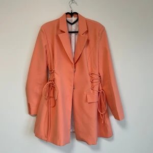 Korallfärgad kavaj med knyt - Snygg korallfärgad kavaj med knyt i midjan och knappar framtill. Den har en klassisk krage och långa ärmar med dekorativa knappar vid ärmsluten. Perfekt för att ge en färgklick till din outfit!