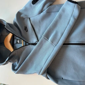 Ljusblå hoodie från Nike - Säljer en stilren ljusblå hoodie från Nike med dragkedja och huva. Jackan har svarta detaljer och en ficka med dragkedja på ärmen. Perfekt för en sportig look!