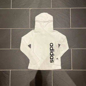 Vit hoodie från Adidas - Säljer en stilren vit hoodie från Adidas med deras logga tryckt i svart på framsidan. Tröjan har långa ärmar och en bekväm huva. Perfekt för en sportig look eller avslappnade dagar.