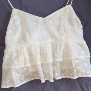 Vit blommig linne - Söt vit linne med blommigt mönster och volanger. Tunna axelband och luftig design, perfekt för varma dagar.