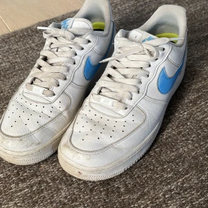 Nike Air Force med blå detaljer - Säljer ett par klassiska Nike Air Force sneakers i vitt med blå swoosh. Skorna har snörning och en bekväm passform. Perfekta för en stilren look. Skorna tvättas innan de skickas