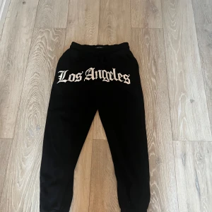 Svarta mjukisbyxor från Black Squad - Svarta mjukisbyxor med texten 'Los Angeles' i vitt på framsidan. Byxorna har en elastisk midja och är perfekta för en avslappnad stil. Tillverkade i ett mjukt material för extra komfort.