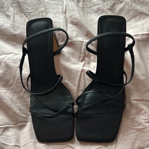 Svarta klackskor från H&M storlek 39 - Eleganta svarta klackskor från H&M med tunna remmar och fyrkantig tå. Perfekta för en stilren look. Använda på fest 1 gång