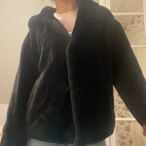Svart teddyjacka med huva ZARA - Jättemysig svart teddyjacka med huva från Zara Basic, perfekt för kyliga dagar. Jackan har en avslappnad passform och stängs med knappar framtill. Skön och håller värmen!! Ett liitet hål i ärmen på baksidan så syns eller känns knappt!! Annars mycket bra skick!
