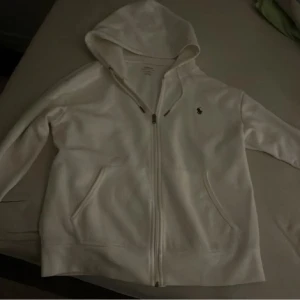 Vit hoodie från Ralph Lauren - Säljer en stilren vit hoodie från Ralph Lauren med dragkedja och huva. Den har en liten broderad logga på bröstet och praktiska fickor framtill. Perfekt för en avslappnad look.