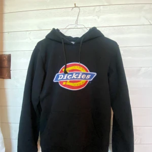 Svart hoodie från Dickies - Säljer en svart hoodie från Dickies med deras ikoniska logga i rött, gult och blått på framsidan. Tröjan har en klassisk passform med långa ärmar och en bekväm huva med dragsko. Perfekt för en avslappnad stil. Ända defekten är att ena plastdelen har lossnat på snöret 