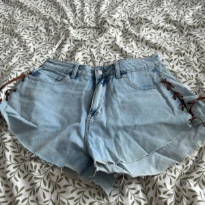 Ljusblå jeansshorts med snörning - Snygga ljusblå jeansshorts med fransiga kanter och cool snörning i sidorna. Perfekta för sommardagar och ger en avslappnad stil. De har en klassisk femficksdesign och knappgylf.