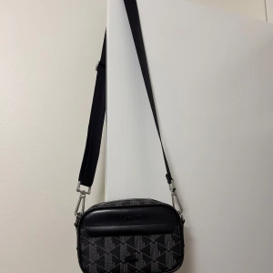 Svart axelväska från Lacoste - Snygg svart axelväska från Lacoste med ett geometriskt mönster. Väskan har en justerbar axelrem och en dragkedja upptill. Perfekt för att bära dina dagliga nödvändigheter med stil.