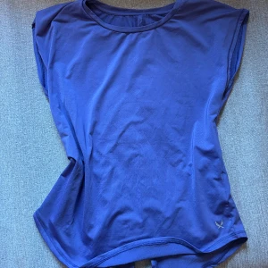 Blå träningstopp från SOC - Säljer en blå träningstopp från SOC. Toppen är nästan ärmlös då det är väldigt korta armar men ej linne och tillverkad i ett lätt och andningsbart material, perfekt för träning. Den har en rund halsringning och en diskret logga på framsidan. Perfekt för aktiva dagar! Andra bilden visar rättvisans färg