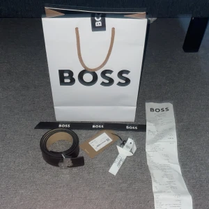 Svart bälte från Boss - Snyggt svart bälte från Boss med en stilren silverfärgad spänne. Perfekt för att ge en elegant touch till din outfit. Bältet är tillverkat i skinn och har en klassisk design som passar till många olika stilar går även att vända på bältet så du får den ljus bruna sidan utåt