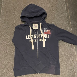 Mörkblå hoodie från Lexington - Säljer en mörkblå hoodie från Lexington med dragkedja och tryck framtill. Tröjan har en broderad amerikansk flagga på bröstet, tröjan har på två ställen lite sämre färg men inget man ser tycker jag, derför går priset ner lite 🙌