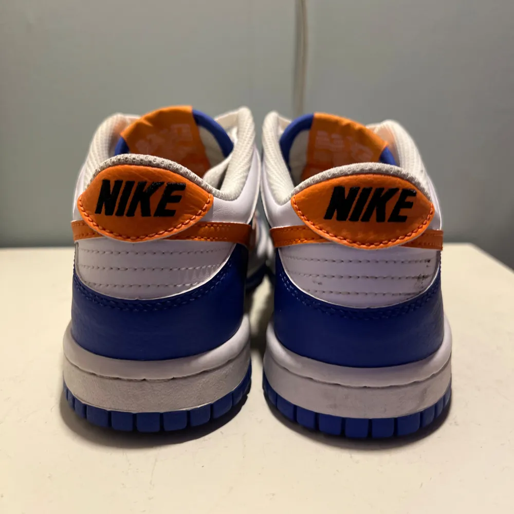 Snygga Nike Dunk sneakers i en fräsch färgkombination av blått och vitt med en iögonfallande orange swoosh. Skorna har klassisk snörning och en bekväm passform, perfekt för en sportig look.. Kengät.