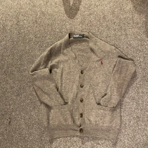  Stickad cardigan från Polo Ralph Lauren - Säljer en stilren grå kofta från Polo Ralph Lauren med knappar framtill och en broderad logga på bröstet. Perfekt för våren! Tröjan är S men skulle säga att den möjligt vis passar XS